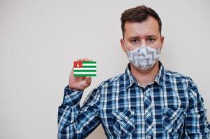 Man in checkered shirt show Abkhazia flag card in hand, wear protect mask isolated on white background. Europe countries Coronavirus concept. photo