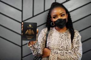 African american woman wearing black face mask show Dominica passport in hand. Coronavirus in America country, border closure and quarantine, virus outbreak concept. photo