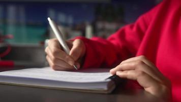una joven con una sudadera rosa brillante se sienta en el escritorio escribiendo en un cuaderno con un bolígrafo