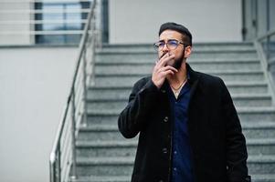 empresario del medio oriente usa abrigo negro y camisa azul, anteojos contra el edificio de oficinas fumando cigarrillos. foto
