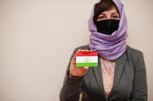Portrait of young muslim woman wearing formal wear, protect face mask and hijab head scarf, hold Tajikistan flag card against isolated background. Coronavirus country concept. photo