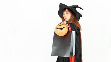 linda chica asiática hermosa con cabello negro y naranja en vestido negro y sombrero de bruja está sosteniendo bolsas de compras, mirando a la cámara y sonriendo, sobre fondo blanco. concepto de halloween foto