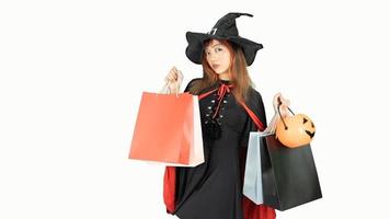 Beautiful girl in black dress and witch hat is holding shopping bags, looking at camera and smiling, on background white. Halloween concept photo