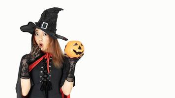 hermosa mujer con cabello negro y naranja en vestido negro y sombrero de bruja sostiene calabaza con emoción wow. concepto de halloween foto