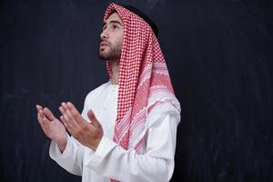 arabian man making traditional prayer to God, keeps hands in praying gesture in front of black chalkboard photo