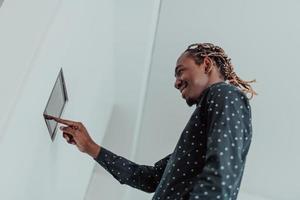 Smiling African American man using modern smart home system, controller on wall, positive young man switching temperature on thermostat or activating security alarm in apartment photo