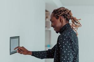 Smiling African American man using modern smart home system, controller on wall, positive young man switching temperature on thermostat or activating security alarm in apartment photo