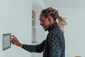 Smiling African American man using modern smart home system, controller on wall, positive young man switching temperature on thermostat or activating security alarm in apartment photo