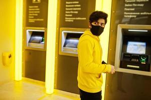 Urban young hipster indian man in a fashionable yellow sweatshirt at bank. Cool south asian guy wear hoodie and black face protect mask, during new normal, withdraws cash at atm. photo