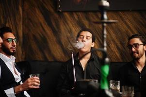 Group of handsome retro well-dressed man gangsters spend time at club, smoking hookah. Multiethnic male bachelor mafia party in restaurant. photo