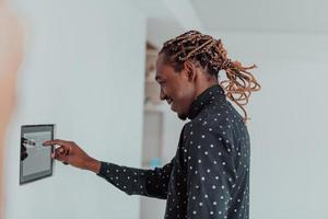 Smiling African American man using modern smart home system, controller on wall, positive young man switching temperature on thermostat or activating security alarm in apartment photo