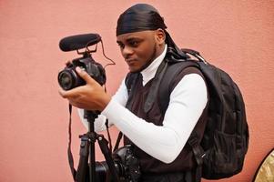 Young professional african american videographer holding professional camera with pro equipment. Afro cameraman wearing black duraq making a videos. photo