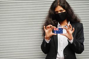 mujer asiática con ropa formal y máscara protectora negra sostiene la bandera de australia a mano contra un fondo gris. coronavirus en el concepto de país. foto