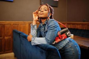 Lovely african american woman with dreadlocks in blue stylish jeans jacket at cafe. Beautiful cool fashionable black young girl indoor. photo
