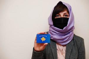 retrato de una joven musulmana que usa ropa formal, protege la máscara facial y el pañuelo en la cabeza del hiyab, sostiene la tarjeta de la bandera de kosovo contra un fondo aislado. concepto de país coronavirus. foto