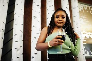 retrato de una atractiva joven afroamericana vestida con suéter verde y jeans negros posan en el restaurante con una copa de vino tinto a mano.