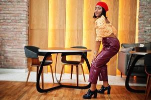 Elegant afro american woman in red french beret, big gold neck chain polka dot blouse and leather pants pose indoor. photo