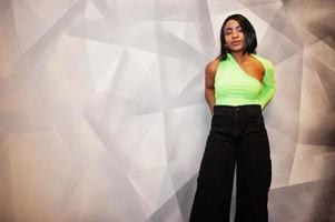 mujer afroamericana de moda con un top verde claro y pantalones negros posan contra una pared gris moderna. foto