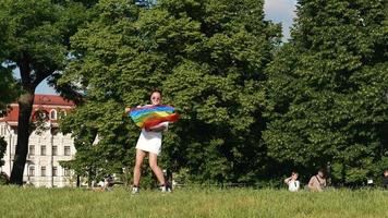 Young woman in white with sunglasses and top knots holds Pride flag and waves it in the wind in a park video