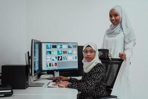 Friends at the office two young Afro American modern Muslim businesswomen wearing scarf in creative bright office workplace with a big screen photo