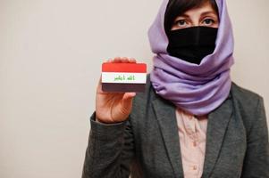 retrato de una joven musulmana que usa ropa formal, protege la máscara facial y el pañuelo de la cabeza del hiyab, sostiene la tarjeta de la bandera de irak contra un fondo aislado. concepto de país coronavirus. foto