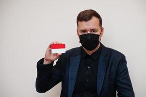 European man wear black formal and protect face mask, hold Monaco flag card isolated on white background. Europe coronavirus Covid country concept. photo