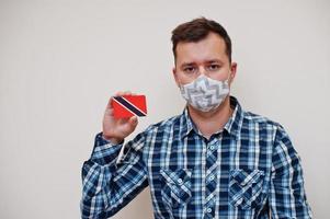 Man in checkered shirt show Trinidad and Tobago flag card in hand, wear protect mask isolated on white background. American countries Coronavirus concept. photo