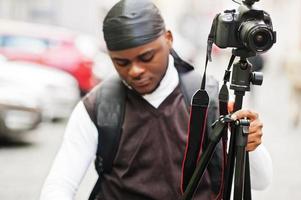 Young professional african american videographer holding professional camera with tripod pro equipment. Afro cameraman wearing black duraq making a videos. photo