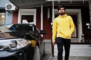 hombre indio joven hipster urbano en una sudadera amarilla de moda. Un tipo genial del sur de Asia usa una sudadera con capucha en el porche de la casa con un auto de negocios negro. foto