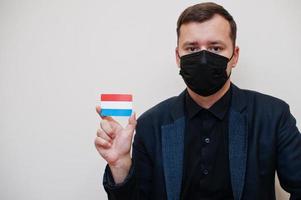 European man wear black formal and protect face mask, hold Luxembourg flag card isolated on white background. Europe coronavirus Covid country concept. photo