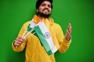 Urban young hipster indian man in a fashionable yellow sweatshirt hold flags of India in hands. Cool south asian guy wear hoodie isolated on green background. photo