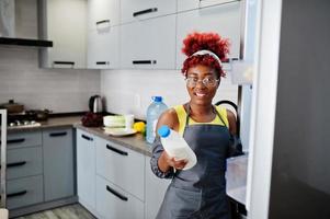 mujer afroamericana en la cocina de casa abre la nevera y tiene una botella de leche. foto