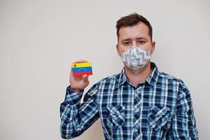 Man in checkered shirt show Venezuela flag card in hand, wear protect mask isolated on white background. American countries Coronavirus concept. photo