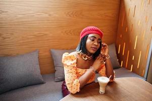 Elegant afro american woman in red french beret, big gold neck chain polka dot blouse pose indoor, sitting at restaurant and drink latte. photo