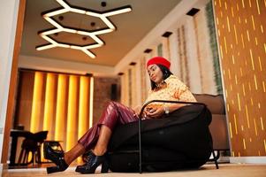 Elegant afro american woman in red french beret, big gold neck chain polka dot blouse and leather pants sit on pouf. photo