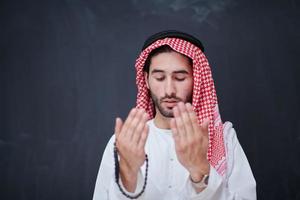 arabian man making traditional prayer to God, keeps hands in praying gesture in front of black chalkboard photo