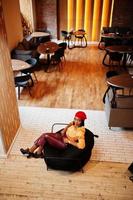 Elegant afro american woman in red french beret, big gold neck chain polka dot blouse and leather pants sit on pouf. photo