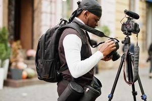 Young professional african american videographer holding professional camera with tripod pro equipment. Afro cameraman wearing black duraq making a videos. photo