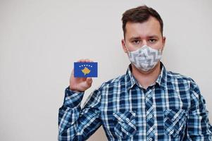 el hombre con camisa a cuadros muestra la tarjeta de la bandera de kosovo en la mano, usa una máscara protectora aislada en el fondo blanco. concepto de coronavirus de los países de europa. foto