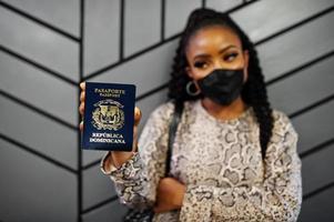 African american woman wearing black face mask show Dominican Republic passport in hand. Coronavirus in America country, border closure and quarantine, virus outbreak concept. photo