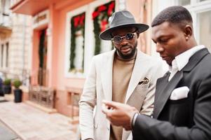 Two fashion black men stand near business car and look at cell phone. Fashionable portrait of african american male models. Wear suit, coat and hat. photo