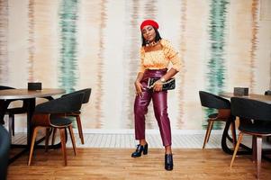 Elegant afro american woman in red french beret, big gold neck chain polka dot blouse and leather pants pose indoor. photo