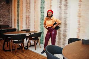 Elegant afro american woman in red french beret, big gold neck chain polka dot blouse and leather pants pose indoor. photo