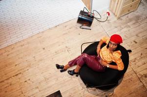 Elegant afro american woman in red french beret, big gold neck chain polka dot blouse and leather pants sit on pouf. photo