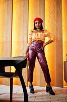 Elegant afro american woman in red french beret, big gold neck chain polka dot blouse and leather pants pose indoor. photo