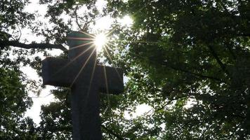 cruz sagrada brillante con rayos de sol brillantes a través del follaje y el cielo brilla una luz de fe espiritual y cristianismo con símbolo de muerte religiosa y resurrección de dioses en la tumba del cementerio al atardecer video