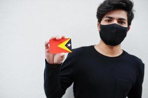 Asian man wear all black with face mask hold East Timor flag in hand isolated on white background. Coronavirus country concept. photo