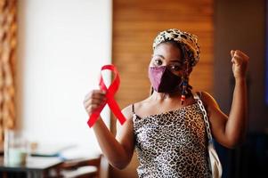 AIDS awareness. Stylish african american woman with dreadlocks afro hair, wear face protect  mask and headdress leopard scarf, hold red ribbon. photo