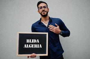 el hombre árabe usa camisa azul y anteojos sostiene una tabla con la inscripción blida algeria. ciudades más grandes en el concepto del mundo islámico. foto