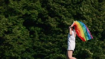 Young woman in white with sunglasses and top knots holds Pride flag and waves it in the wind in front of trees ata park video